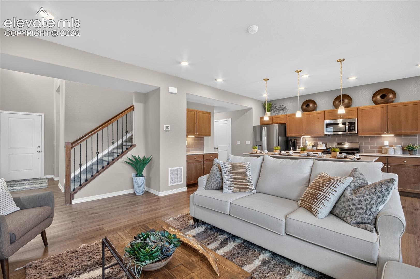 Open-concept layout featuring wood-finish flooring, recessed lighting, a staircase with a stained wooden handrail and metal balusters, and a kitchen with wooden cabinetry, stainless steel appliances, and pendant lighting