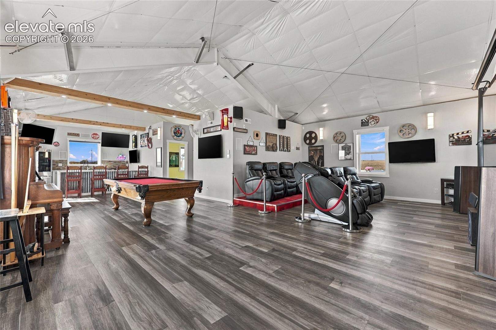 Expansive interior space featuring vaulted ceilings with exposed wood beams, wood-finish flooring, a built-in wet bar, and a raised platform area