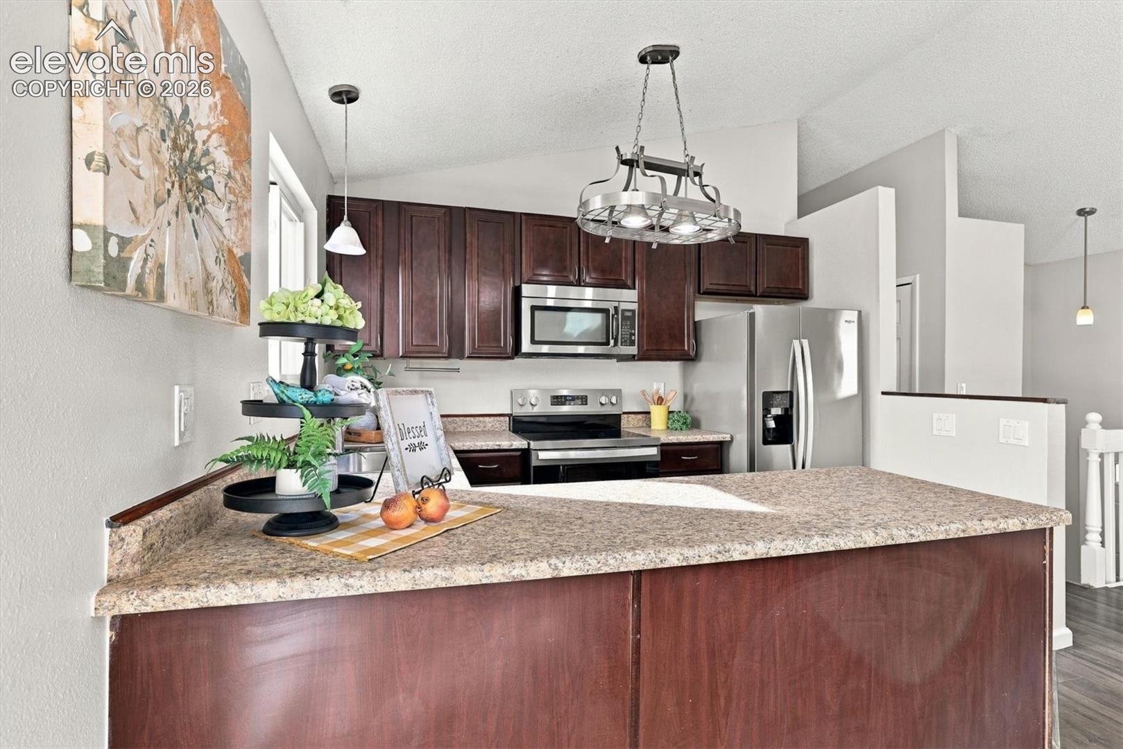 Kitchen featuring dark wood cabinetry, stainless steel appliances, a peninsula with stone-finish countertops, and decorative metal pendant lighting