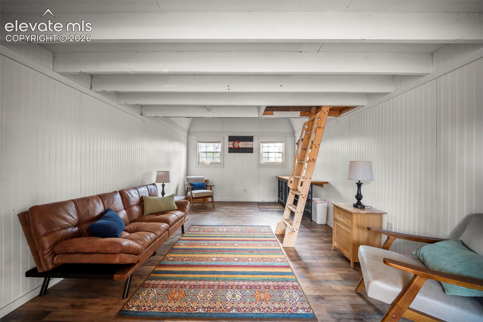 Interior space featuring exposed ceiling beams, vertical wood-panelled walls, wood-finish flooring, two windows with natural lighting, and an integrated wooden loft ladder