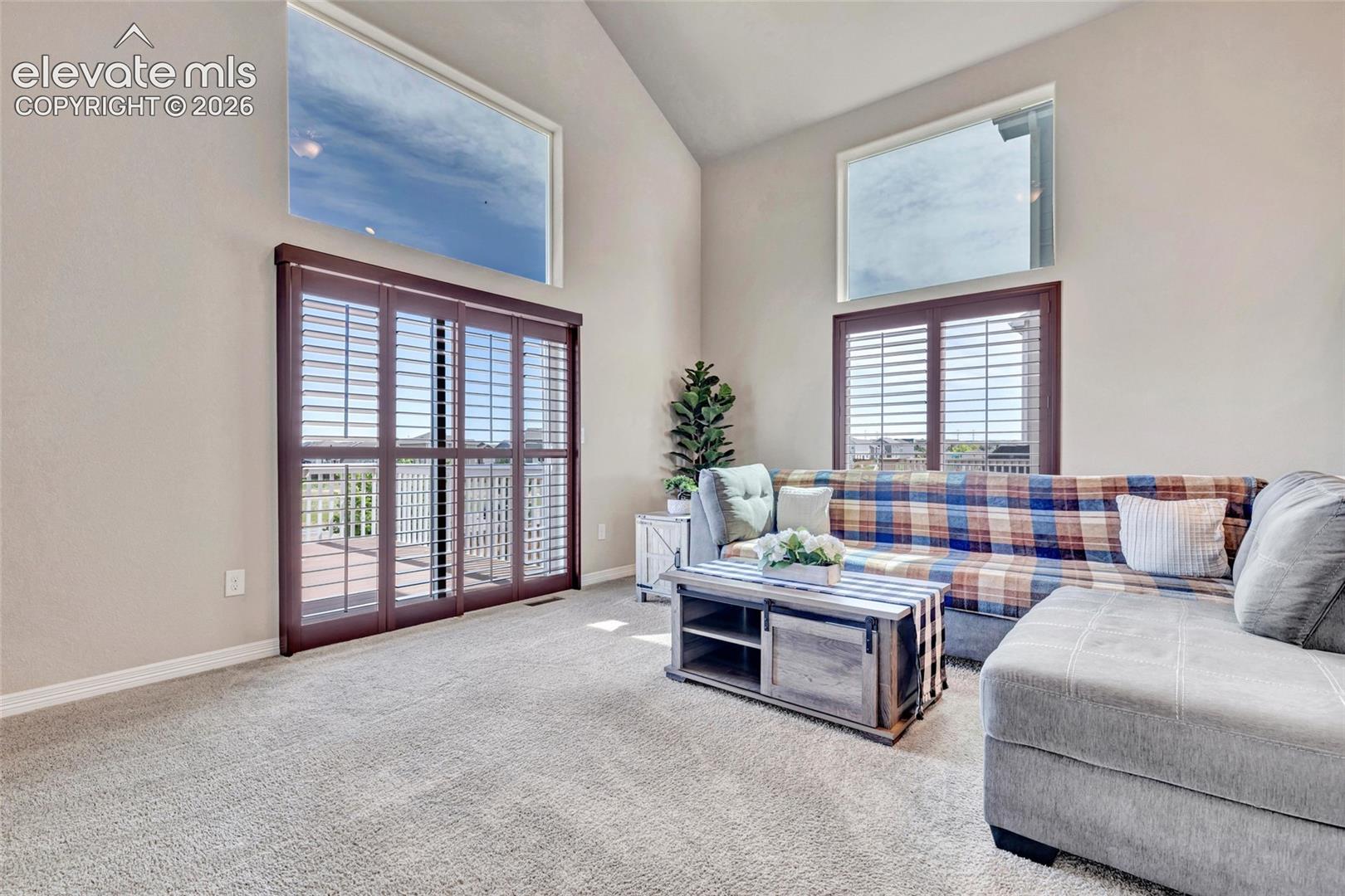 Spacious living area featuring vaulted ceilings, light-toned carpeting, and a sliding glass door with plantation shutters