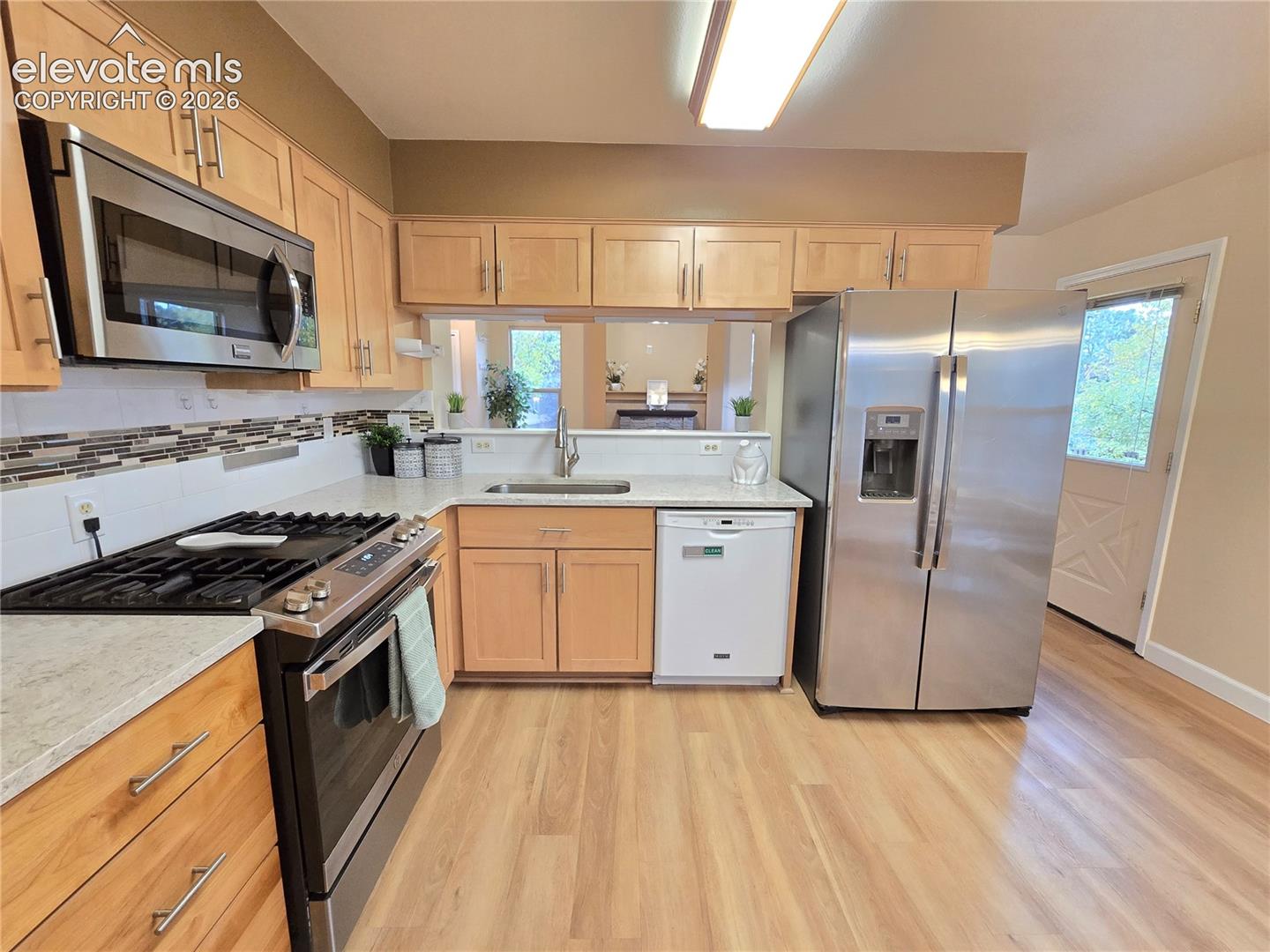 Kitchen featuring light wood cabinetry, stainless steel appliances, a stone-tile backsplash, light-colored countertops, and wood-finish flooring