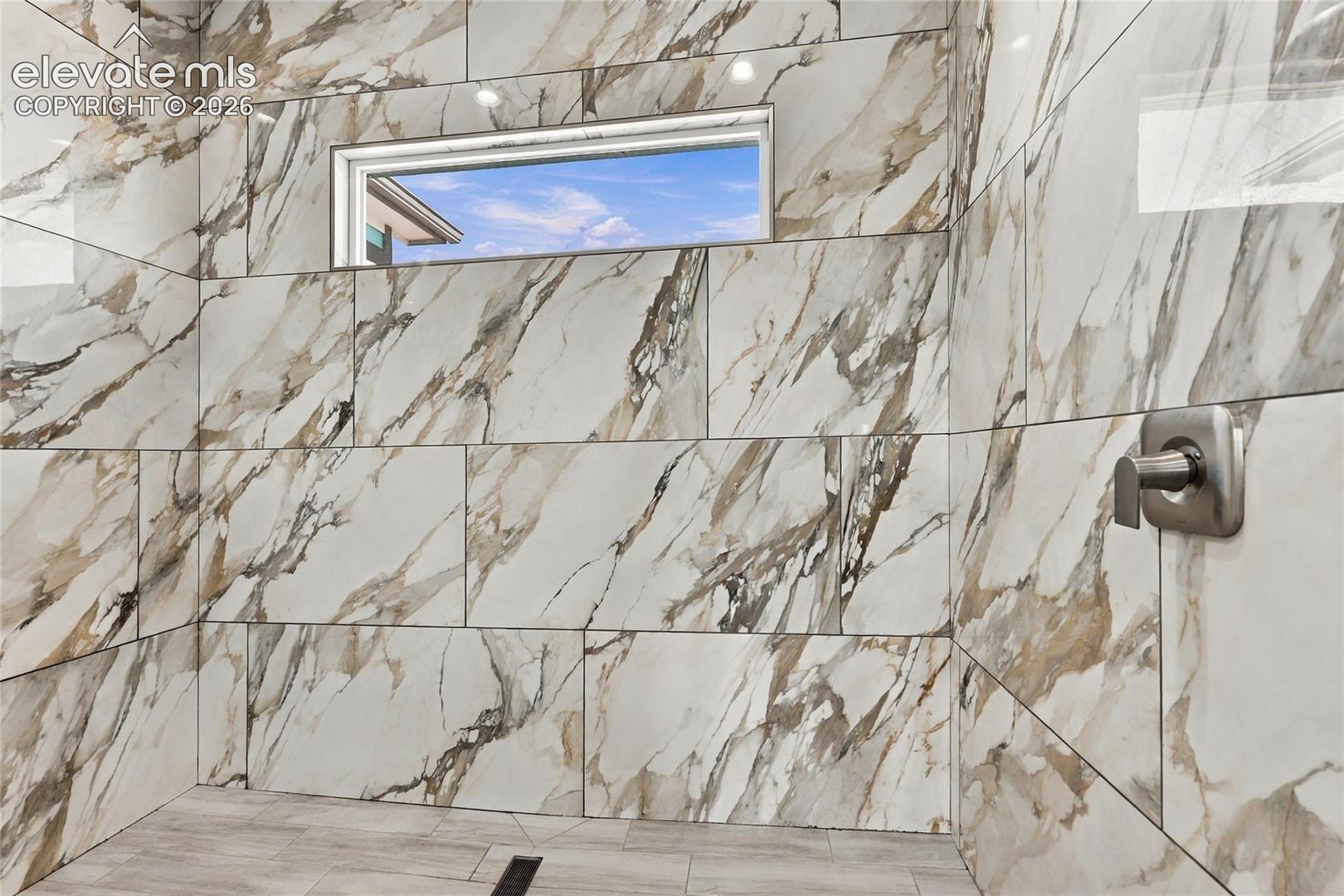 Walk-in shower featuring large-format patterned wall tile, linear floor drain, horizontal transom window, and brushed metallic faucet hardware