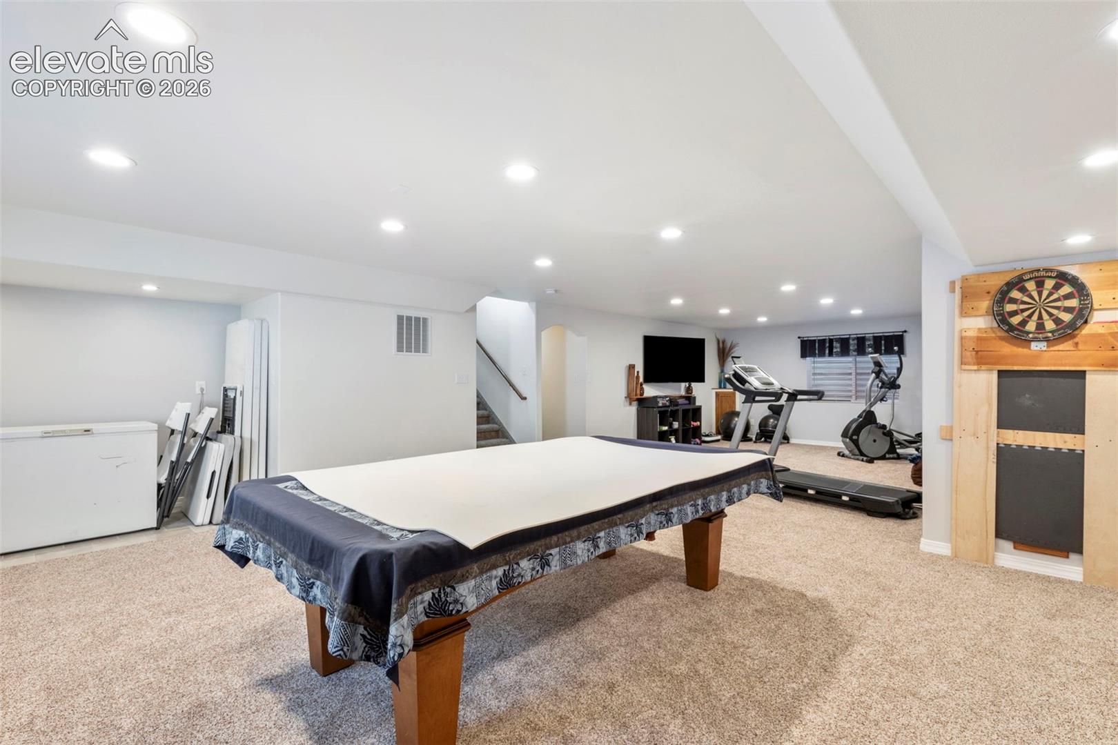 Expansive carpeted area with recessed lighting, a dartboard wall feature, and a staircase