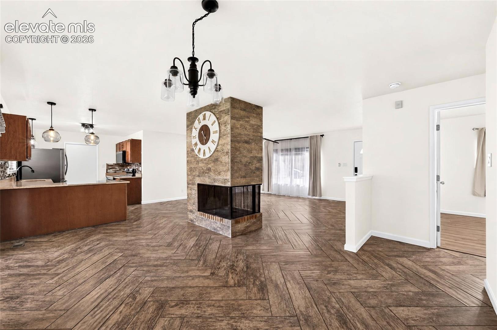 Central stone-clad dual-sided fireplace, herringbone-patterned wood-finish flooring, island kitchen with wood cabinetry, and multi-light hanging fixtures