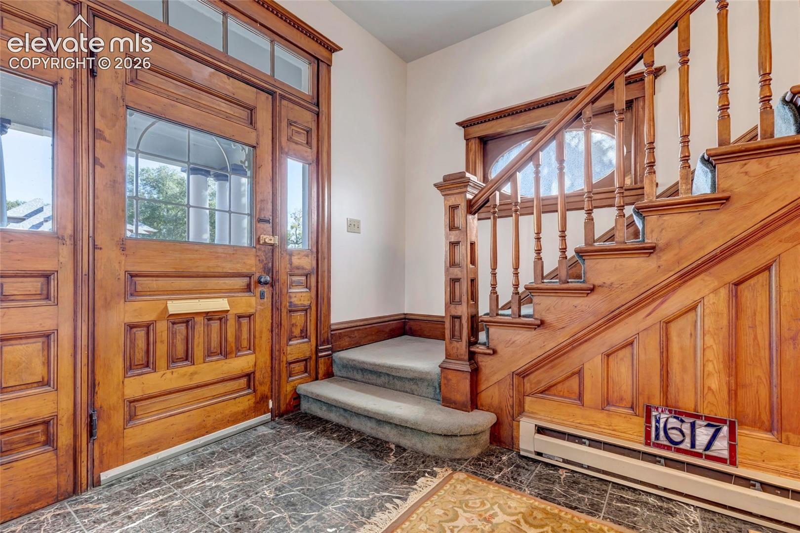 Entryway featuring extensive natural wood paneling, a solid wood paneled front door with a transom window, an ornate wood staircase with decorative balusters, and dark stone flooring