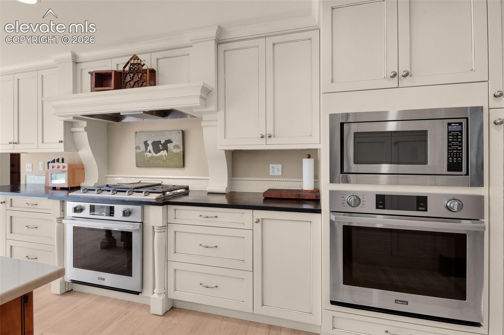 Off-white custom cabinetry, dark solid-surface countertops, built-in stainless steel wall oven and microwave, professional-style gas range, and wood-finish flooring