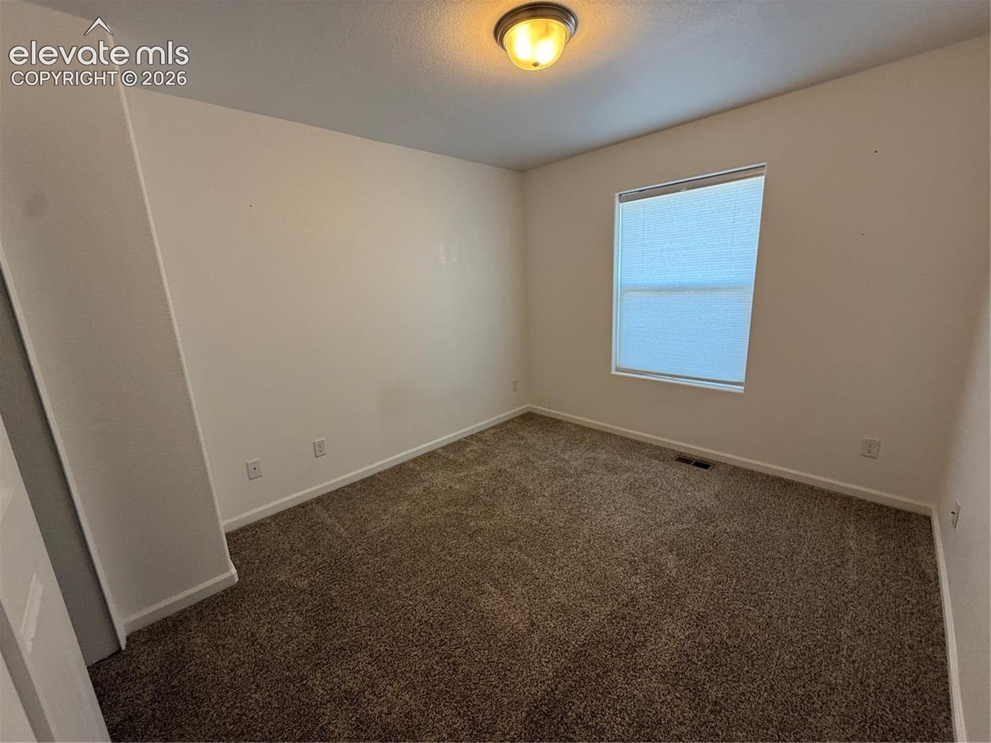 Neutral-toned interior space featuring plush carpeting, soft wall coloring, a single-pane window with a fitted blind, and a flush-mount overhead light fixture