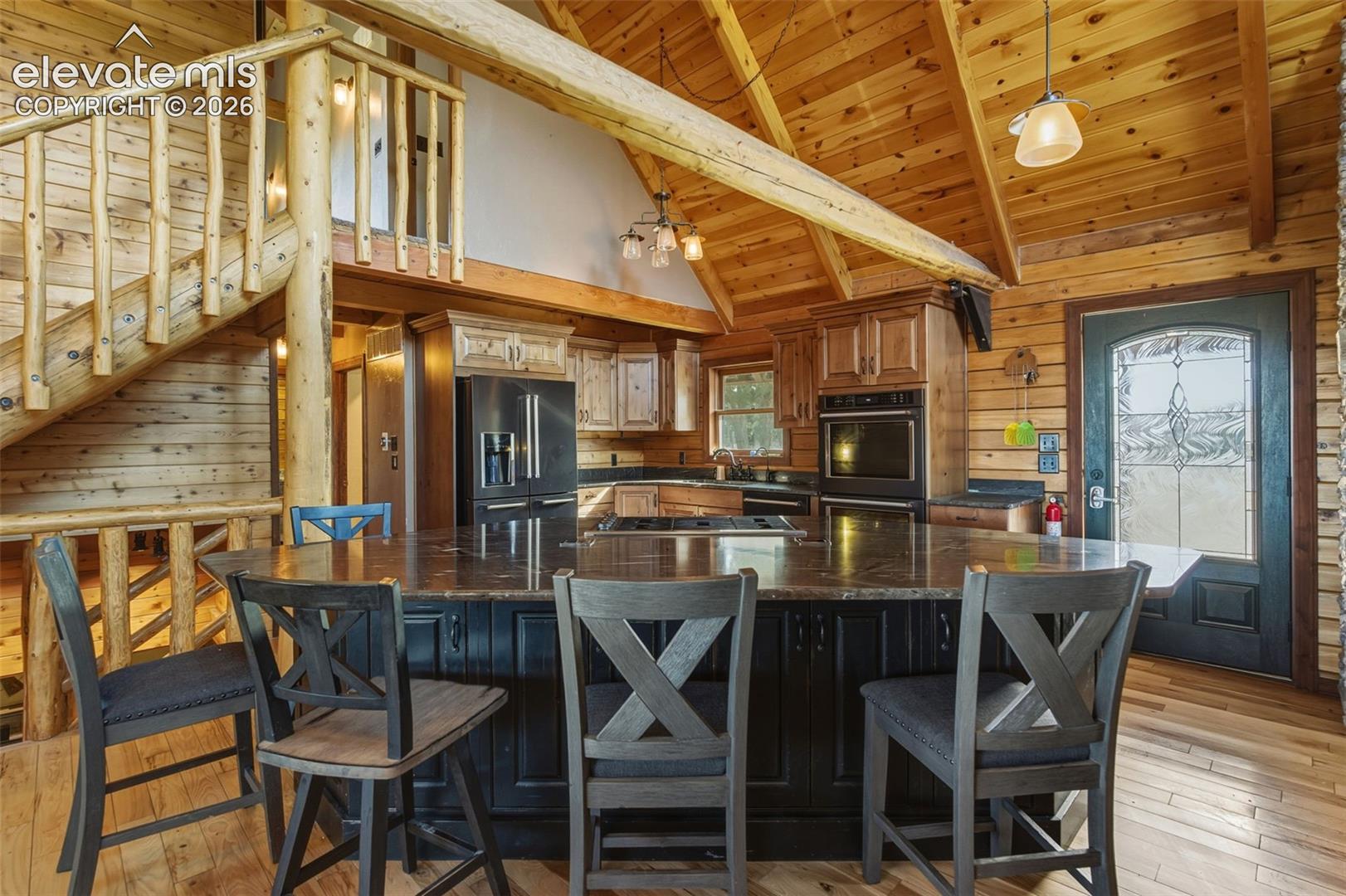 Vaulted wood-paneled ceiling with exposed beams, wood-finish flooring, kitchen island with dark cabinetry and stone countertop, stainless steel refrigerator, and integrated oven