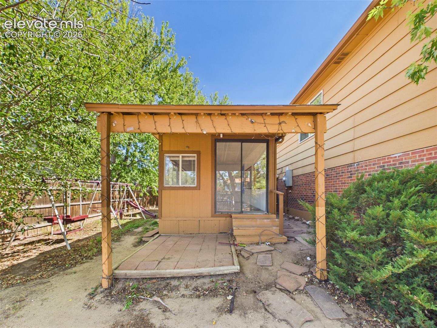 Covered patio area extends alongside the enclosed sunroom, offering a sheltered outdoor space surrounded by mature landscaping.