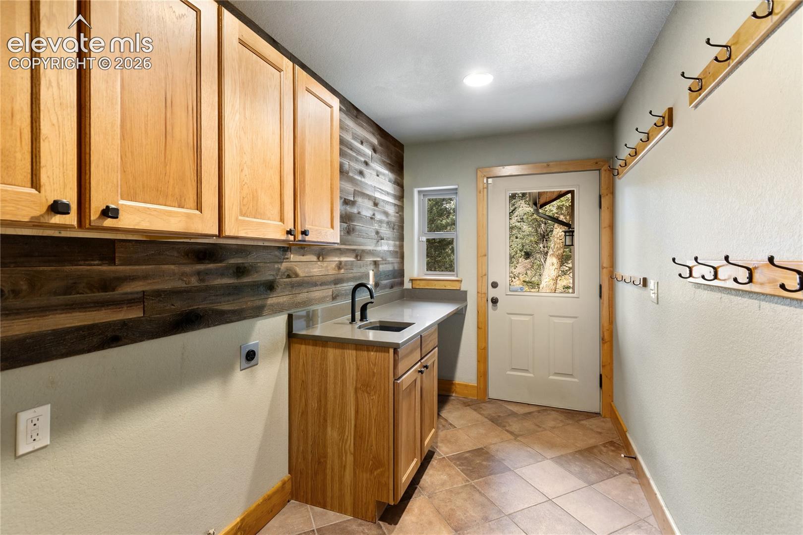 Utility space featuring wood-finish cabinetry, a neutral countertop with an integrated sink, matte black hardware, textured wood-panel wall accents, and ceramic tile flooring