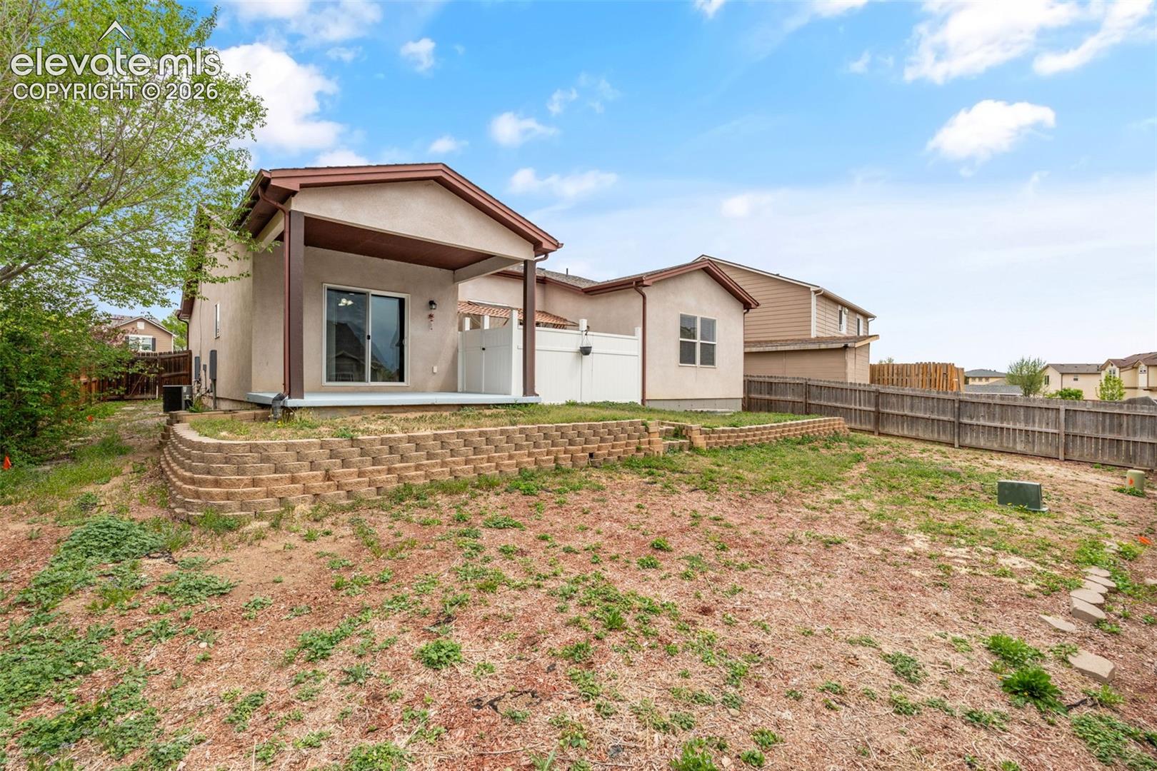Rear exterior featuring a covered patio, tiered decorative block retaining wall, white vinyl privacy fencing, and wood-panel perimeter fencing