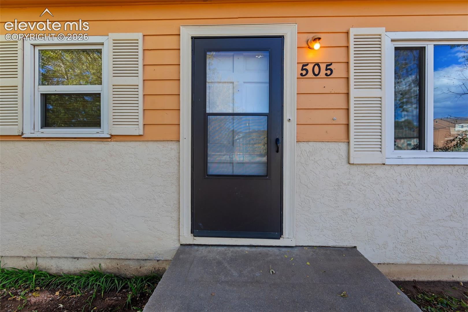 Exterior entrance featuring horizontal lap siding, a textured stucco lower facade, white louvered window shutters, and a concrete stoop