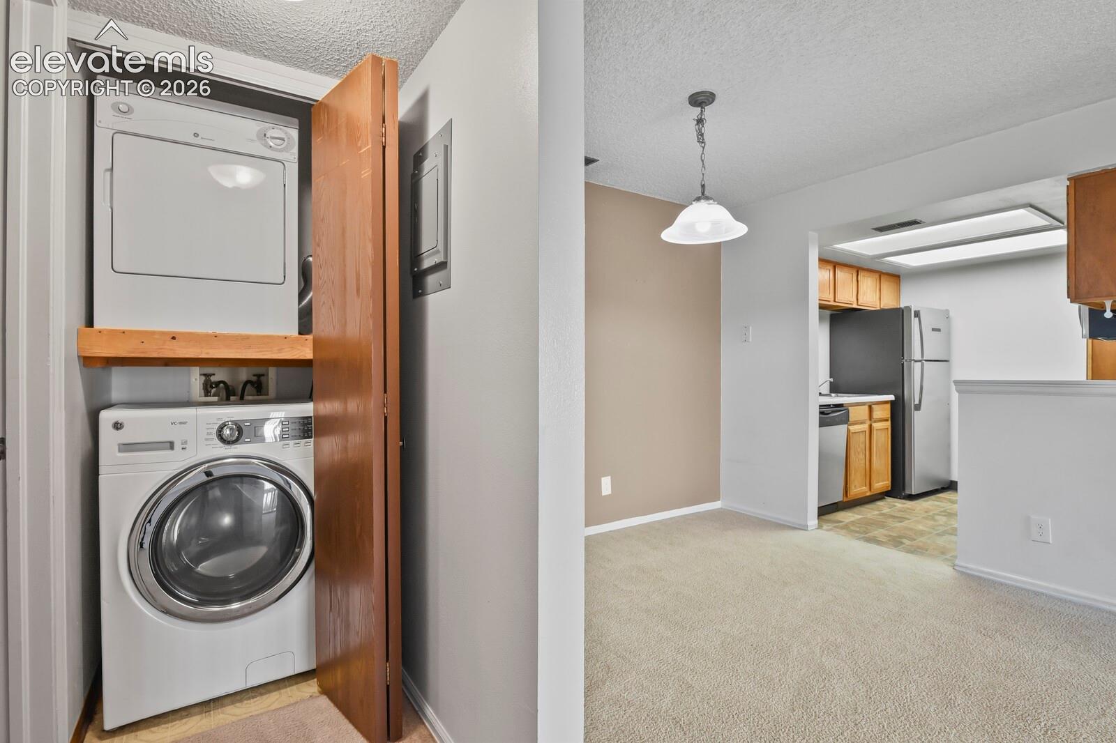 Stacked laundry closet with built-in wooden shelving, neutral carpeting, pendant lighting, and kitchen equipped with wood-finish cabinetry and stainless-steel appliances
