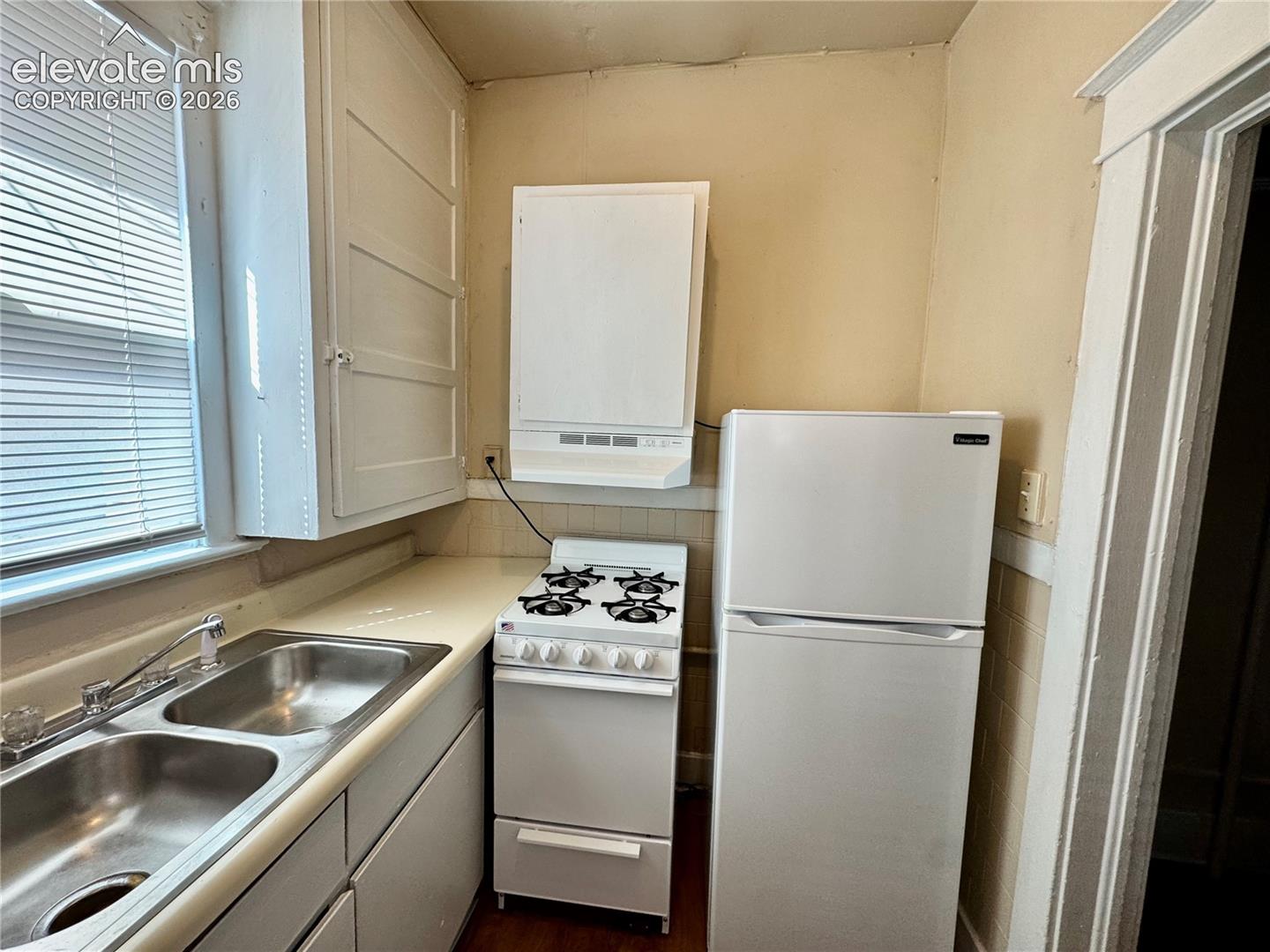 Compact kitchen featuring stainless steel double-basin sink, gas range with four burners, white cabinetry, white refrigerator with freezer compartment, and window with blinds allowing natural light