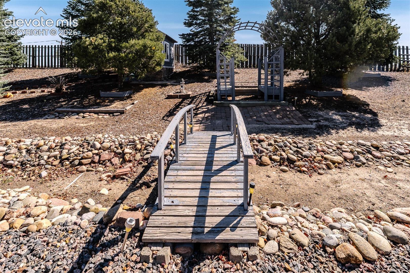 Backyard decorative bridge and arched trellis to a garden shed behind the trees.