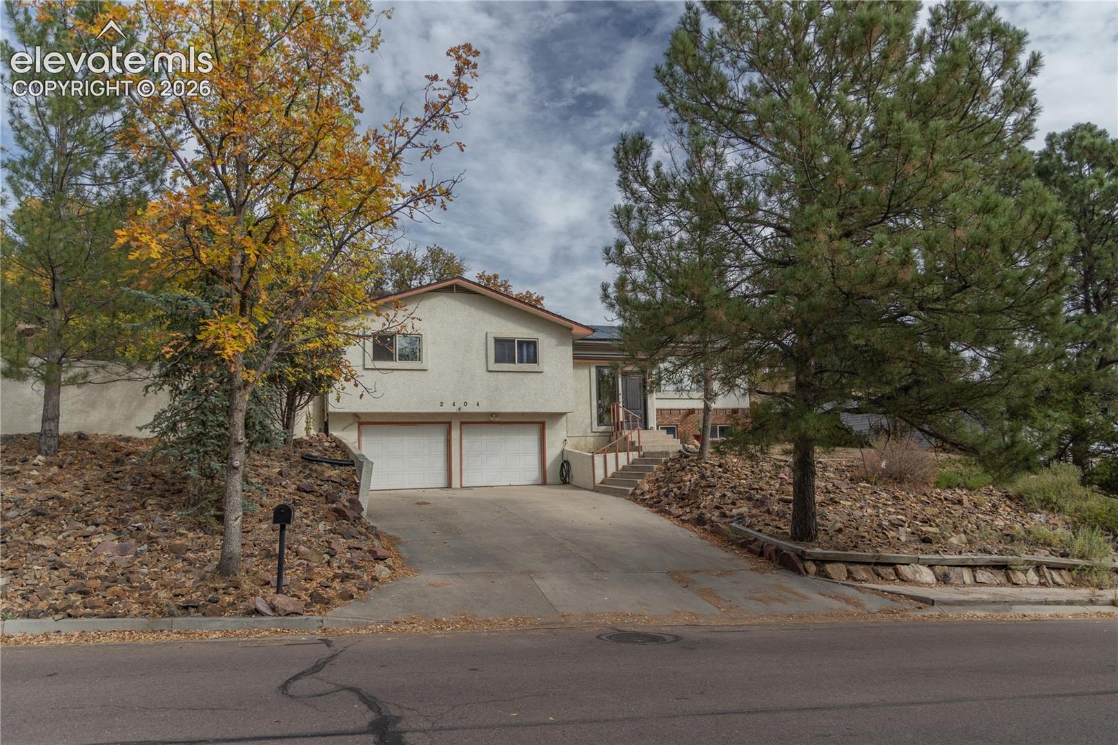 Split-level exterior featuring a stucco facade, attached two-car garage, concrete driveway, and natural rock landscaping with mature evergreen and deciduous trees