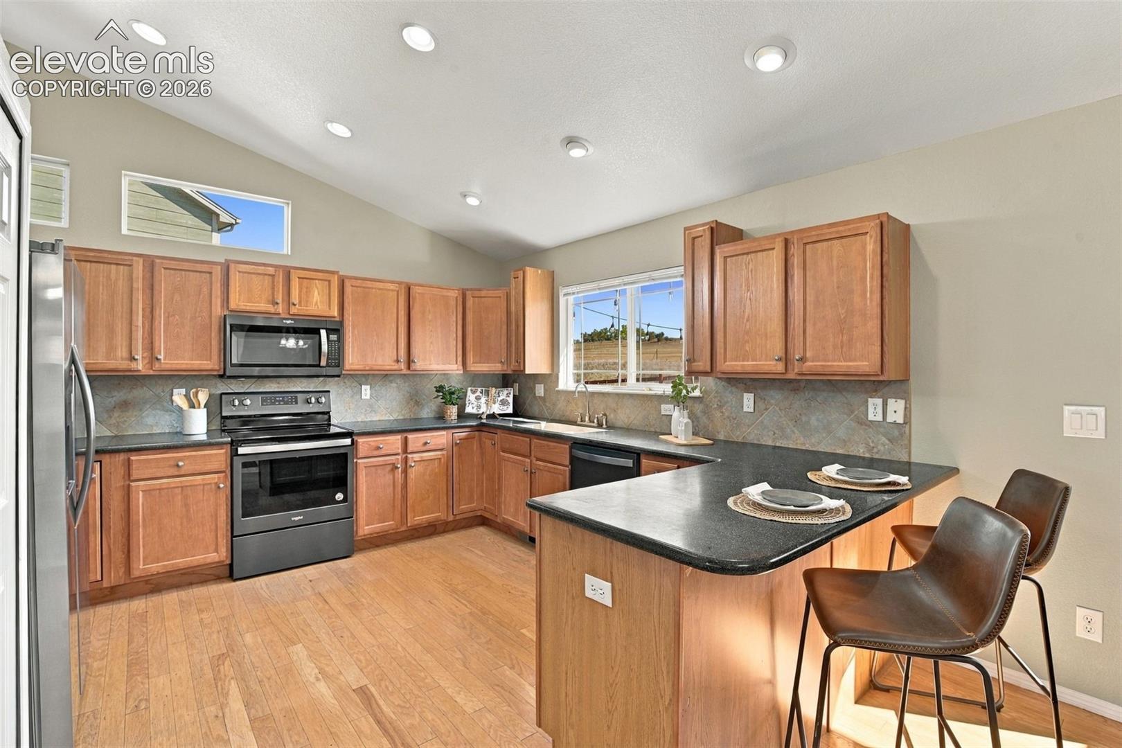 Kitchen with wood-finish flooring, recessed lighting, vaulted ceiling, wood cabinetry, and dark countertops