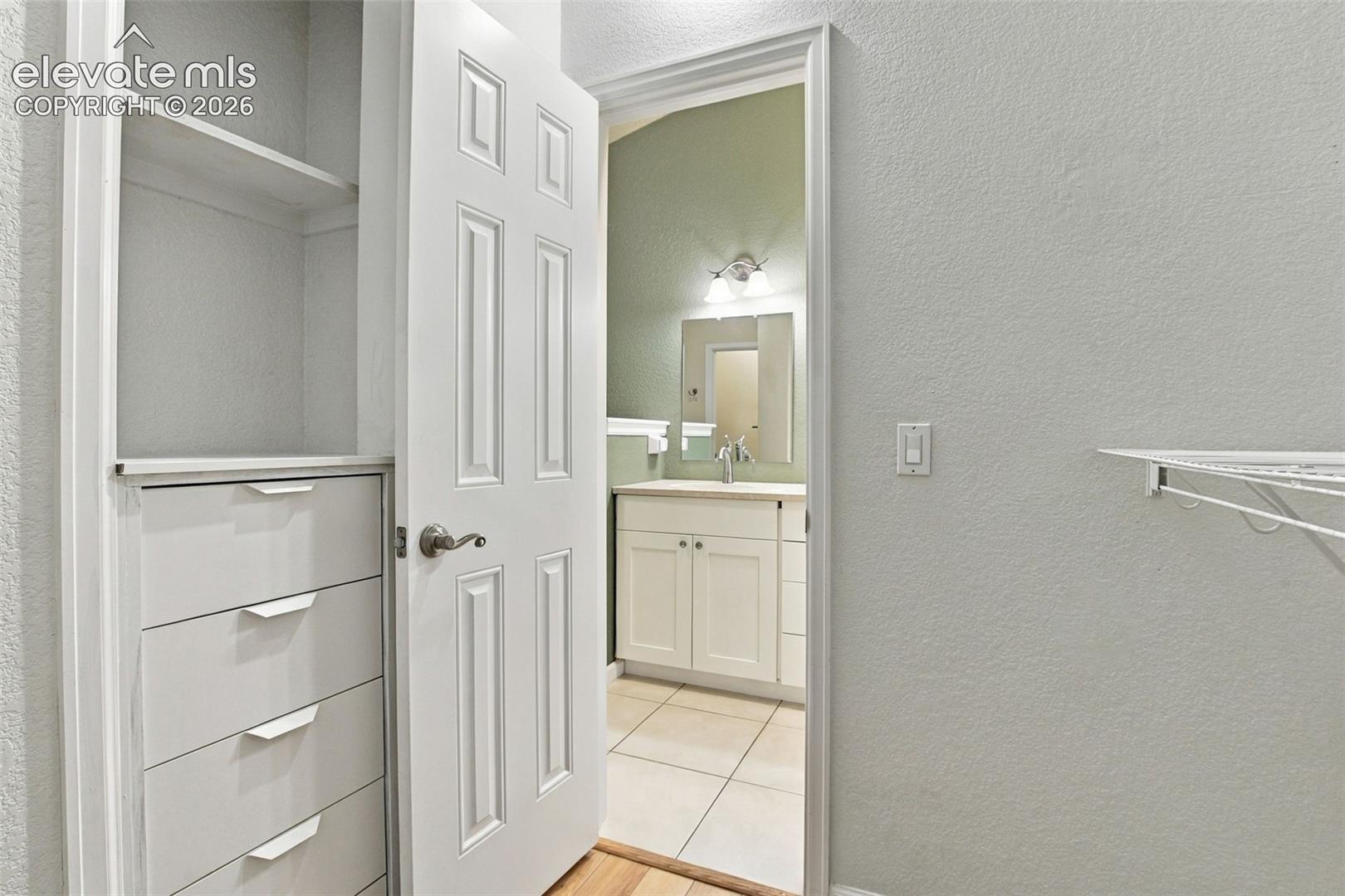 Built-in cabinetry featuring open shelving and four pull-out drawers, white six-panel door, wire shelving, vanity with white shaker-style cabinetry, and tile flooring