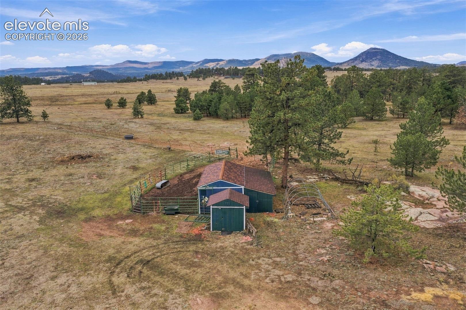 Expansive lot featuring an outbuilding with a shingled roof, fenced enclosure, metal hoop structure, mature pine trees, and a mountain backdrop