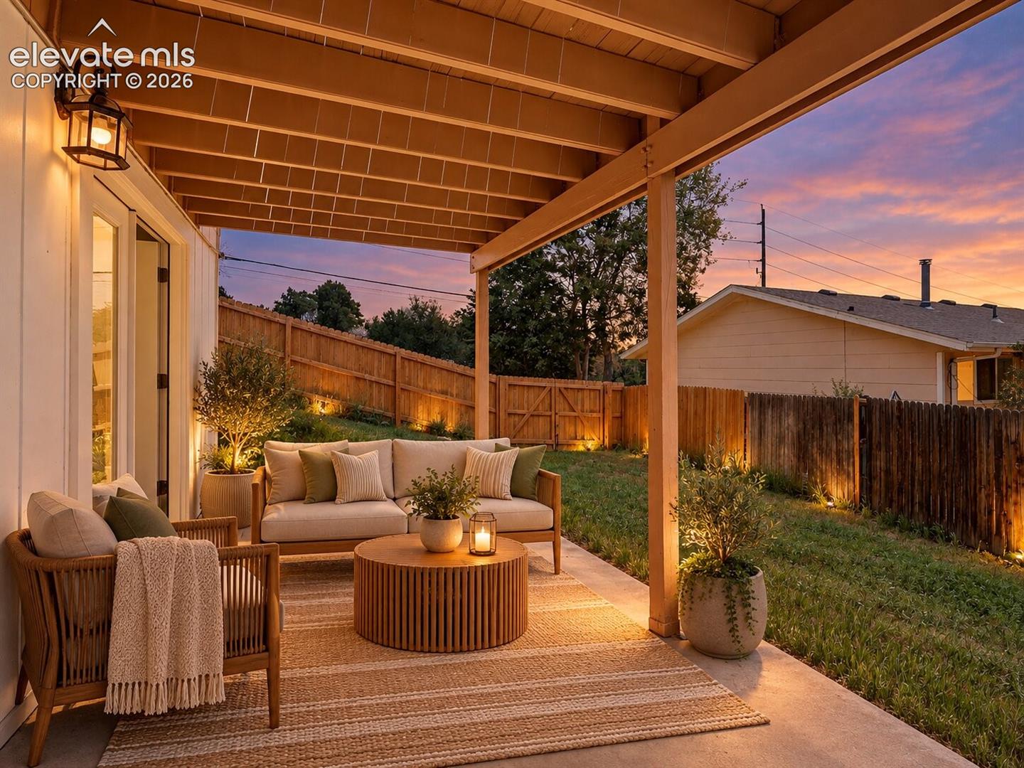 Covered patio featuring an exposed-beam ceiling, wall-mounted lantern-style lighting, and concrete flooring, complemented by an enclosed backyard with wooden privacy fencing. AI-Enhanced – furniture/decor digitally added