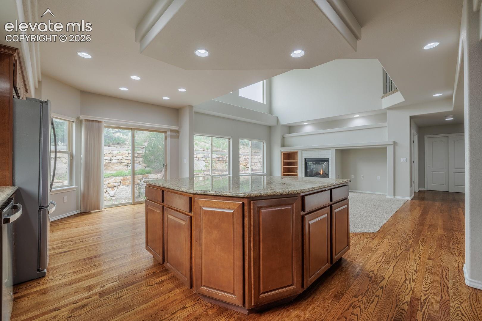 Open-concept kitchen and great room featuring a substantial wood cabinetry island with multi-sided design, wood-finish flooring throughout, recessed and pendant lighting, abundant natural light from multiple large windows, and soaring vaulted ceilings with skylights