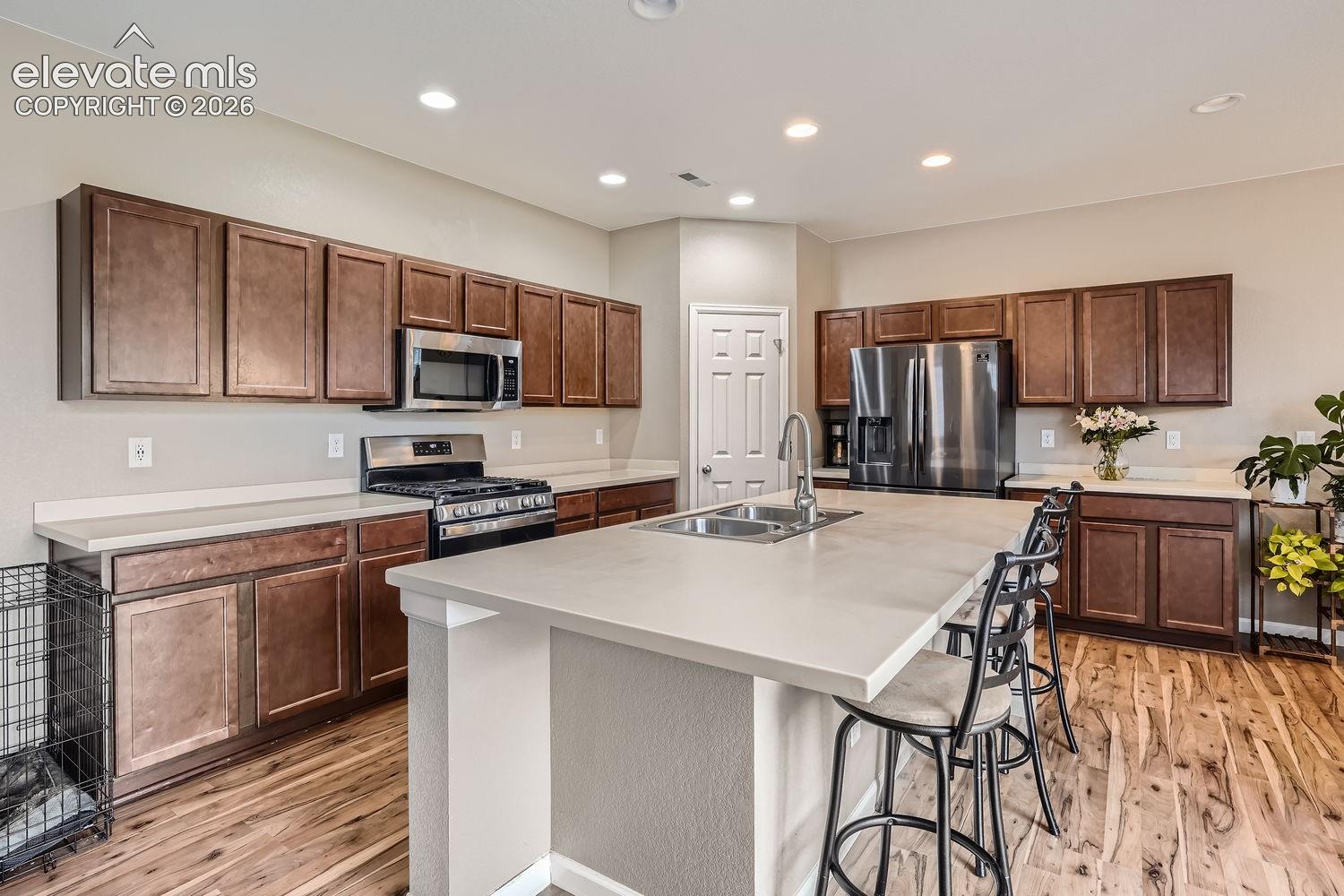 Kitchen featuring stained wood cabinetry, a central island with a double-basin stainless steel sink, solid surface countertops, stainless steel appliances, and wood-finish flooring