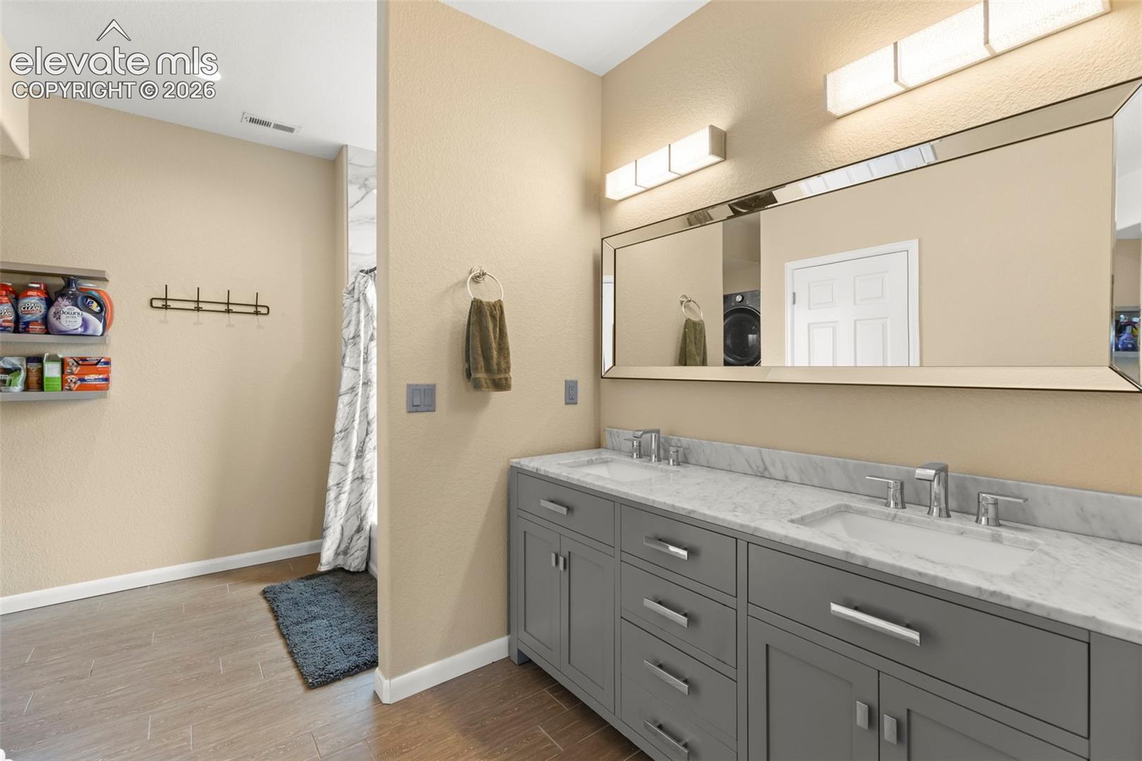 Dual-sink vanity with a stone countertop, cabinetry with metal hardware, a wall-mounted mirror, and wood-finish flooring