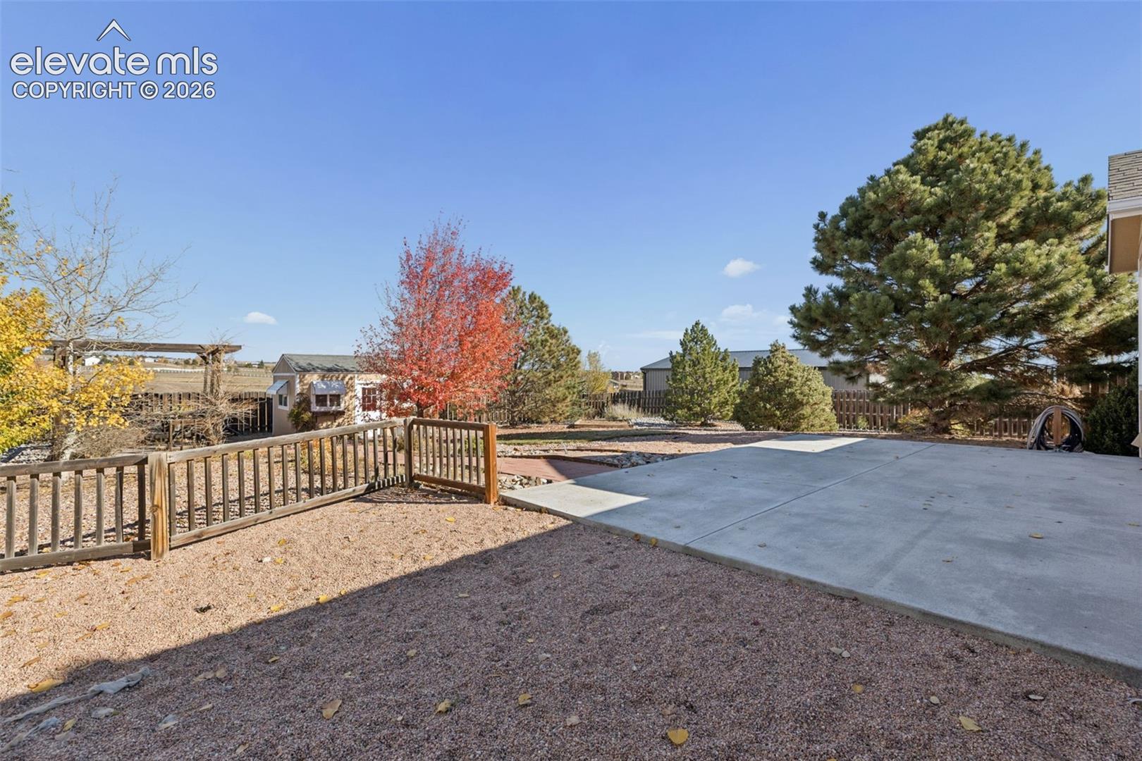 Expansive backyard featuring a concrete patio, wood-slat perimeter fencing, mature evergreen trees, and a decorative wooden pergola
