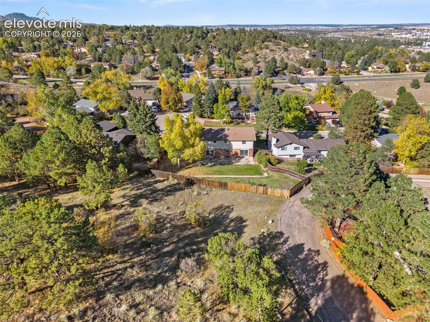 Aerial perspective of a multi-level residence featuring an expansive lot, wood-slat fencing, paved driveway, and established pine trees