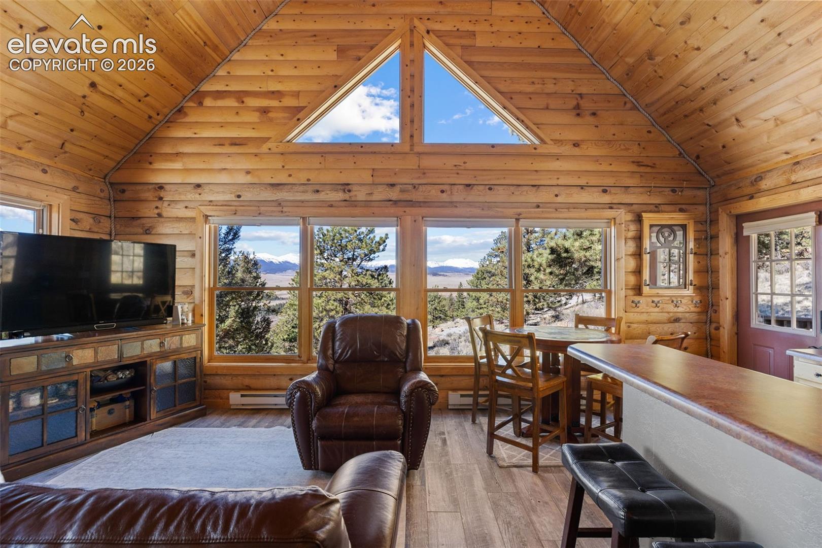 Vaulted wood-paneled ceiling, exposed log walls, large triangle-shaped fixed window, expansive picture windows, and wood-finish flooring