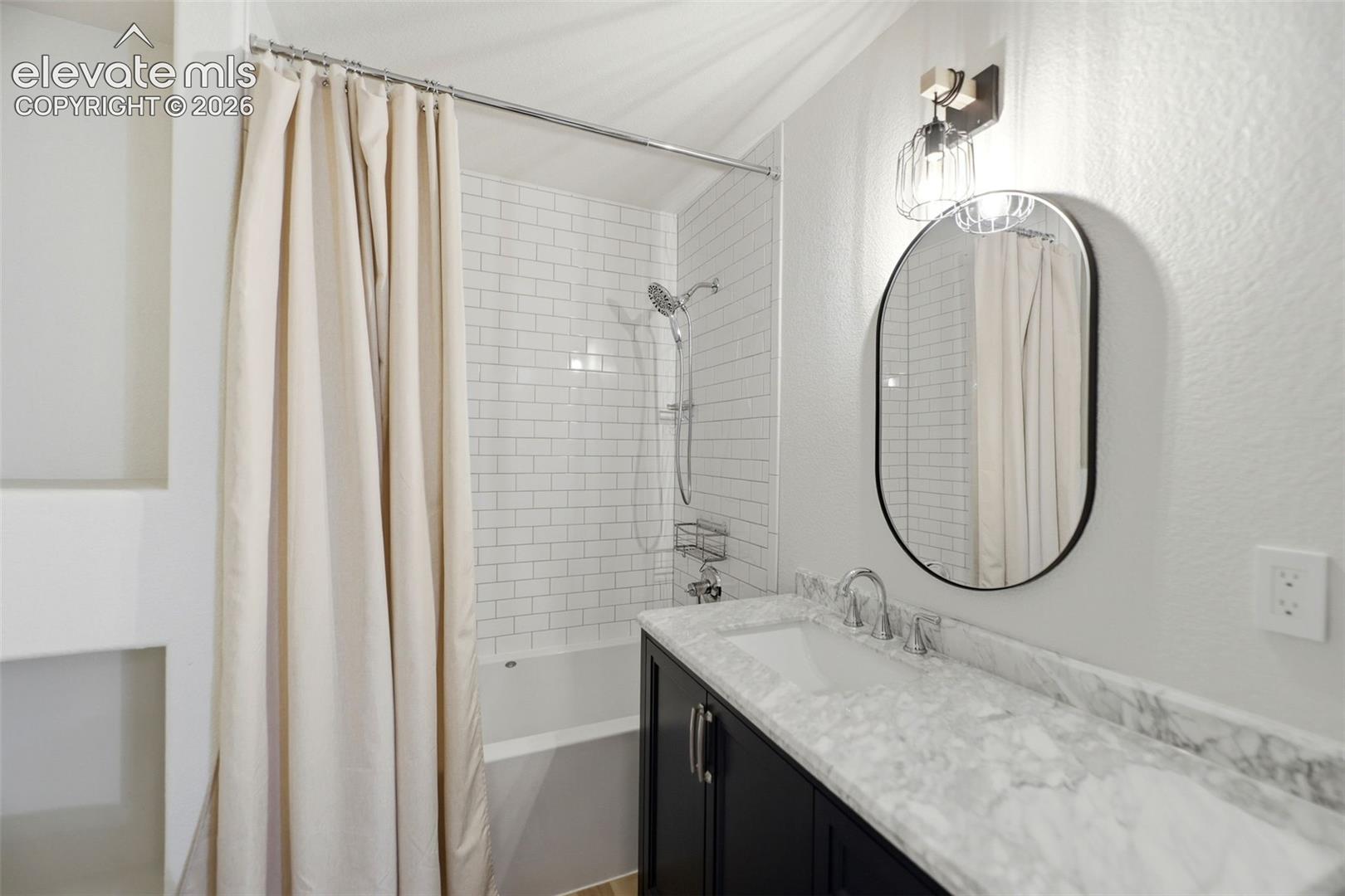 Bathroom featuring a vanity with a white stone countertop, undermount sink, dark cabinetry, and an oval mirror with wall-mounted lighting