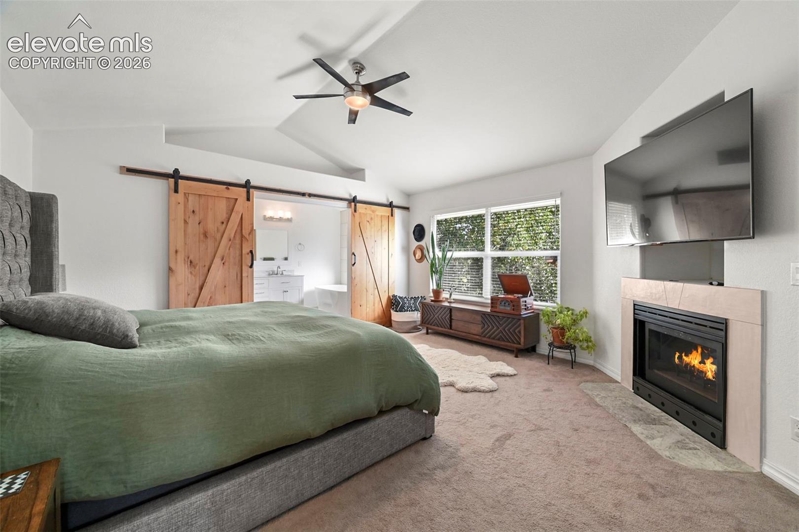 Vaulted ceilings with a ceiling fan, dual sliding barn doors, an integrated fireplace with a tiled surround, and multiple windows