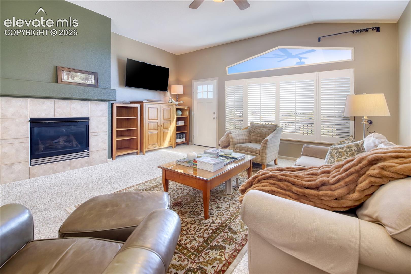 Living area featuring a tile-surround fireplace, built-in wooden cabinetry, vaulted ceilings with an integrated ceiling fan, and large windows fitted with plantation shutters