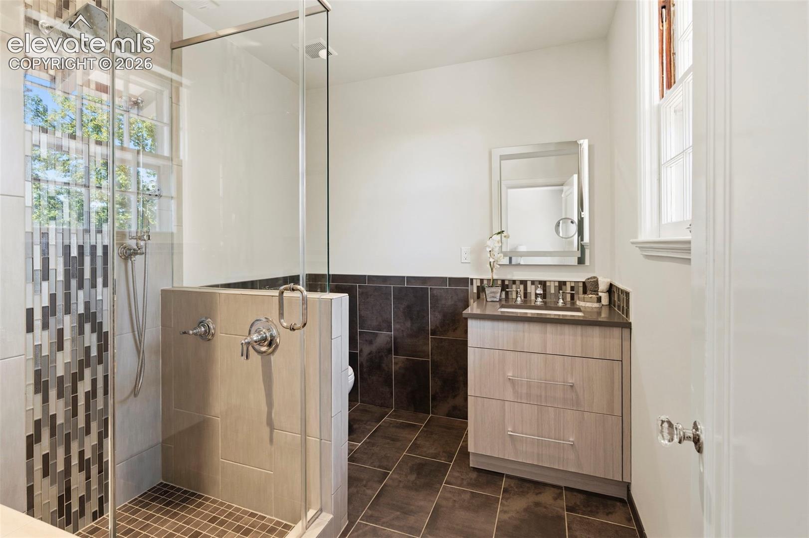 Walk-in shower with a glass enclosure, wood-finish vanity, solid surface countertop, rectangular undermount sink, and tile flooring