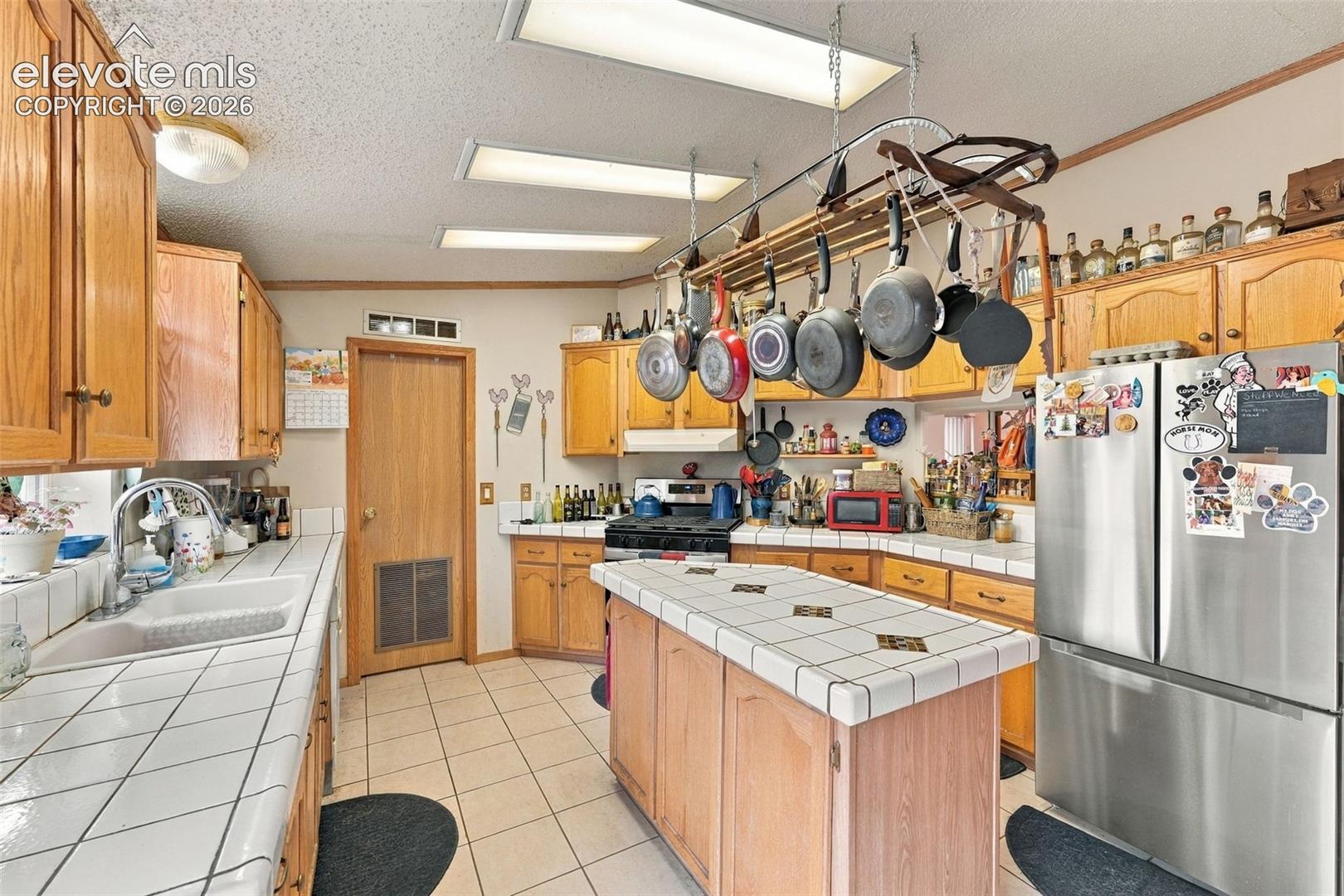 Kitchen featuring wood cabinetry, a central island with tile countertops, tile flooring, stainless steel refrigerator, and recessed lighting