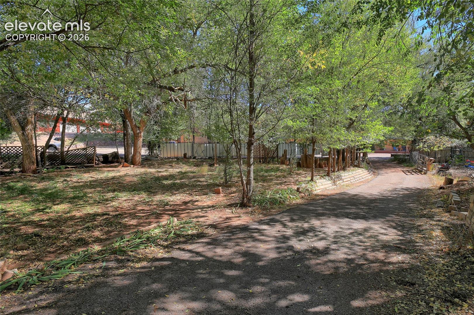 Expansive grounds featuring a paved driveway, mature trees, tiered retaining wall, and wood lattice fencing