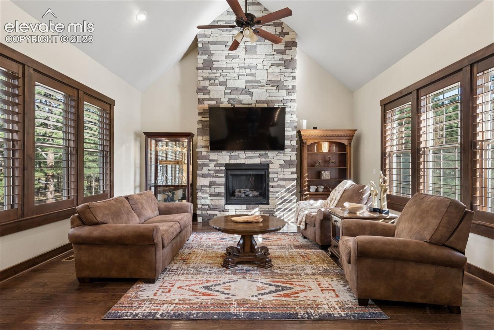 Vaulted ceiling with a central paddle fan, full-height stacked stone fireplace, wood-finish flooring, and multi-pane windows with integrated shutters
