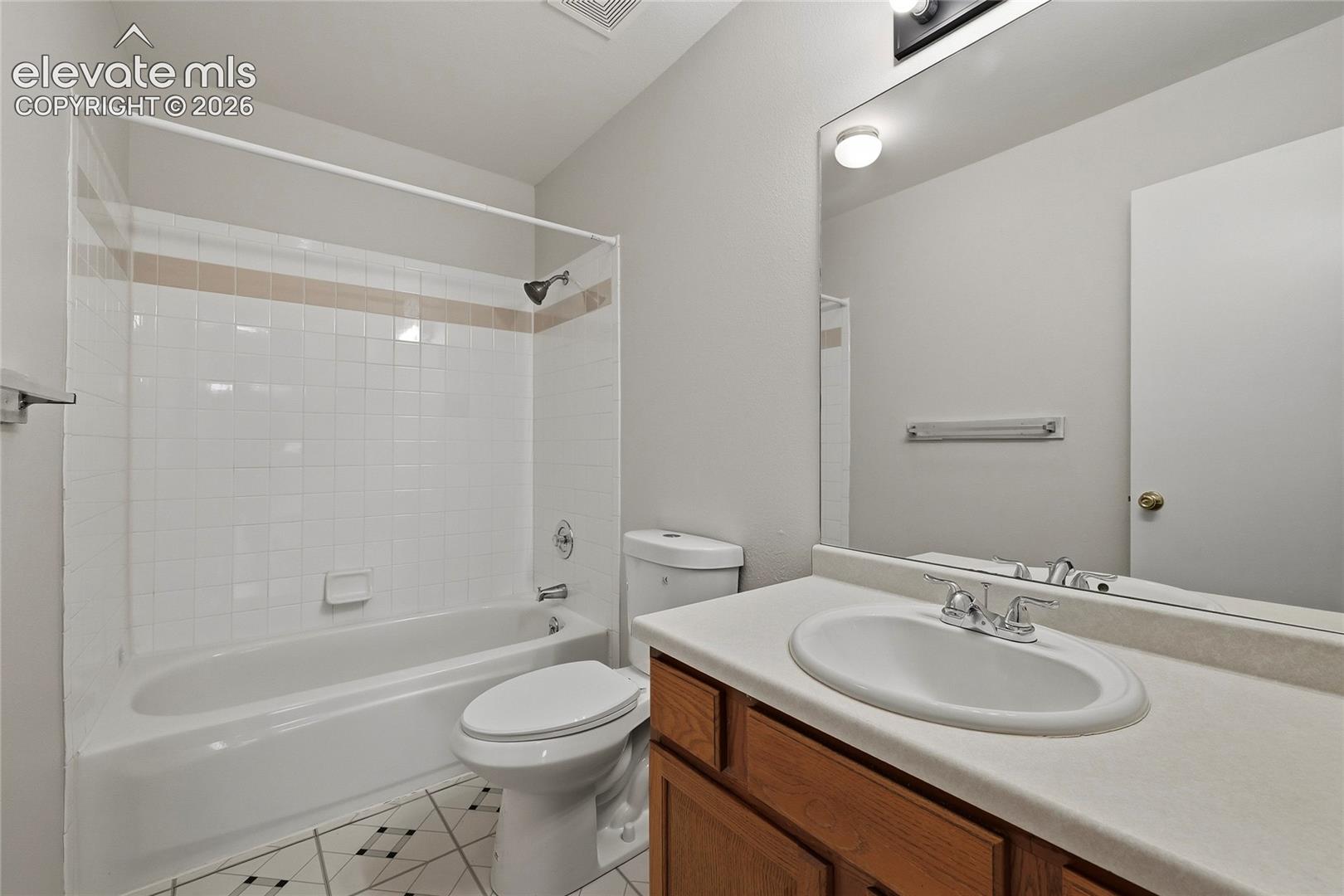 Full-width vanity with integrated sink, large wall-mounted mirror, tub-shower combination with white tile surround, and patterned tile flooring
