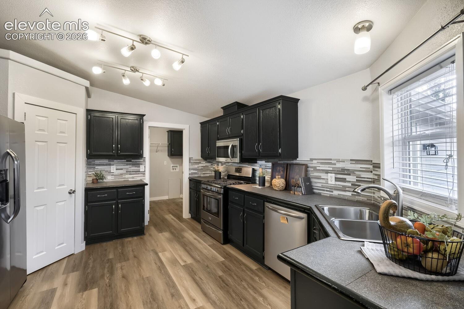 Kitchen featuring black cabinetry, wood-finish flooring, multi-tonal tile backsplash, stainless steel appliances, and dual-track lighting fixtures