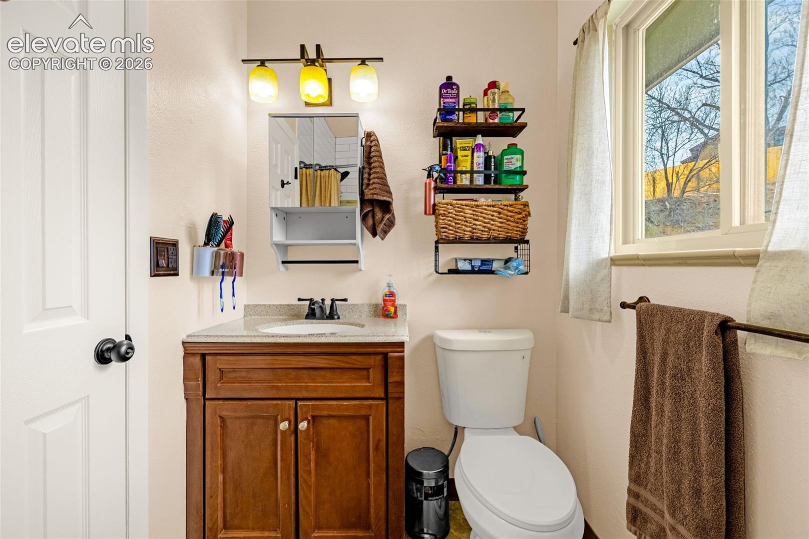 Wood-finish vanity with a stone-composite countertop, drop-in sink, dark-finished hardware, tri-light vanity fixture, and mirrored medicine cabinet