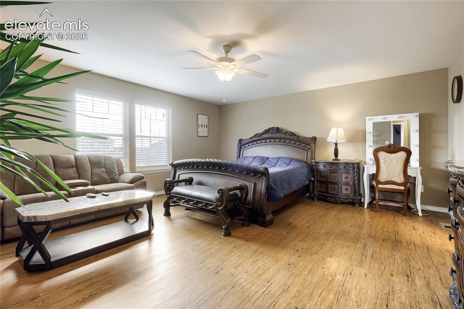 Spacious primary bedroom featuring wood-finish flooring, sitting area, neutral-toned walls, and multiple windows with blinds