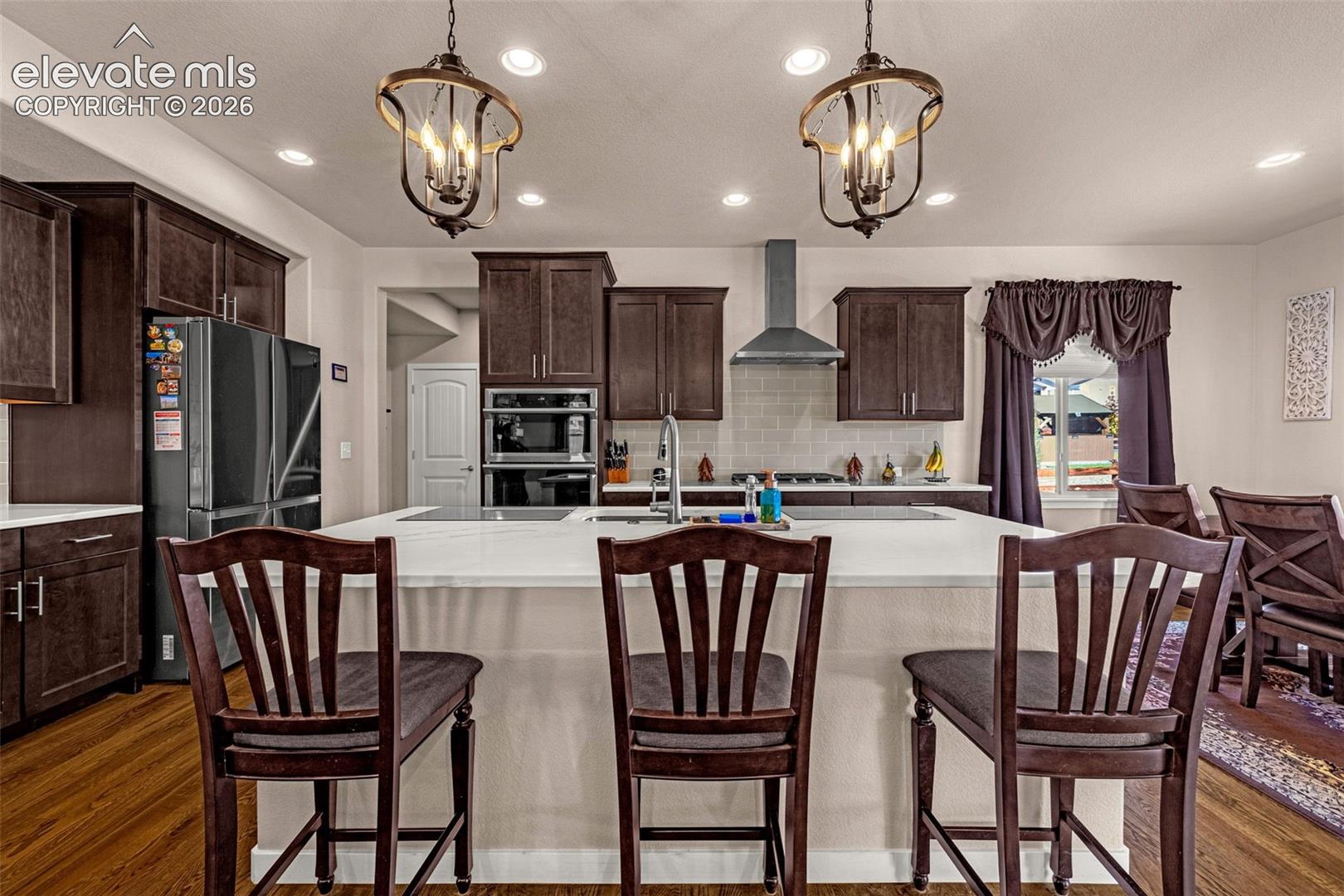 Kitchen featuring dark cabinetry, expansive central island with a quartz countertop, recessed lighting, stainless steel wall-mounted range hood, and wood-finish flooring