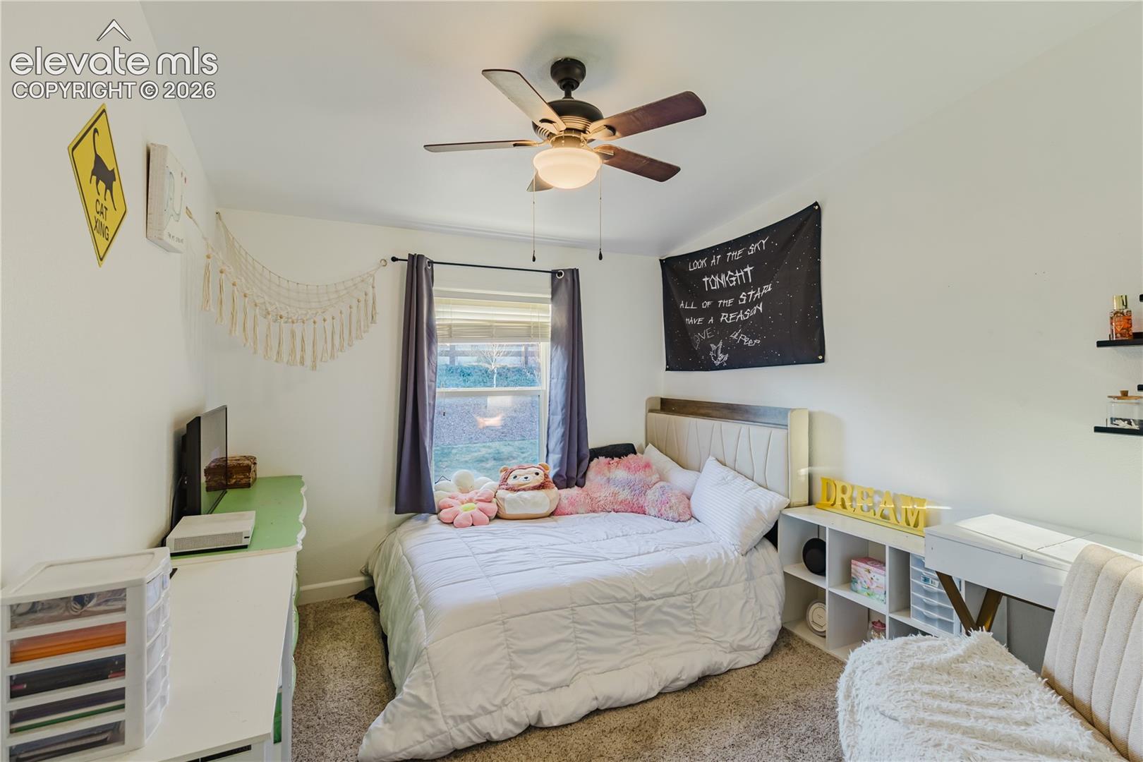 Wall-to-wall carpeting, ceiling fan with integrated lighting, window with dark-toned drapes, and an upholstered headboard