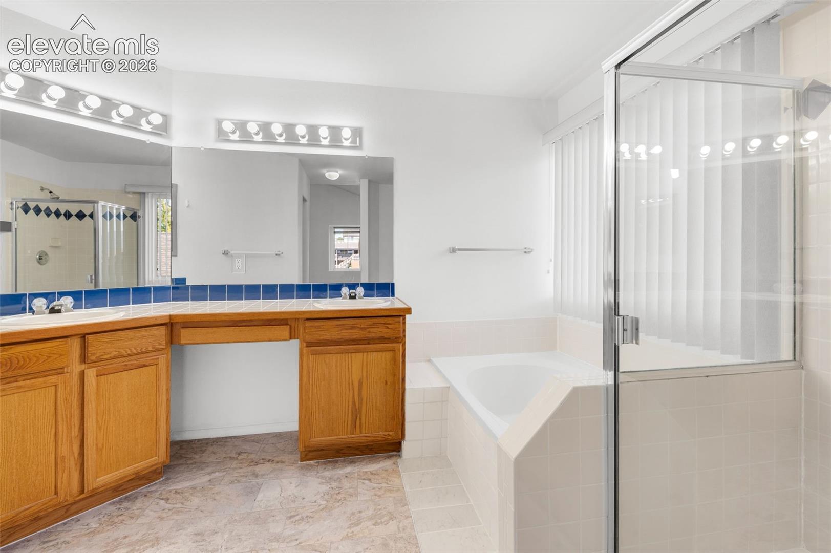 Dual vanity with wood cabinetry, large mirrors, built-in soaking tub, glass-enclosed shower, and light-toned tile flooring