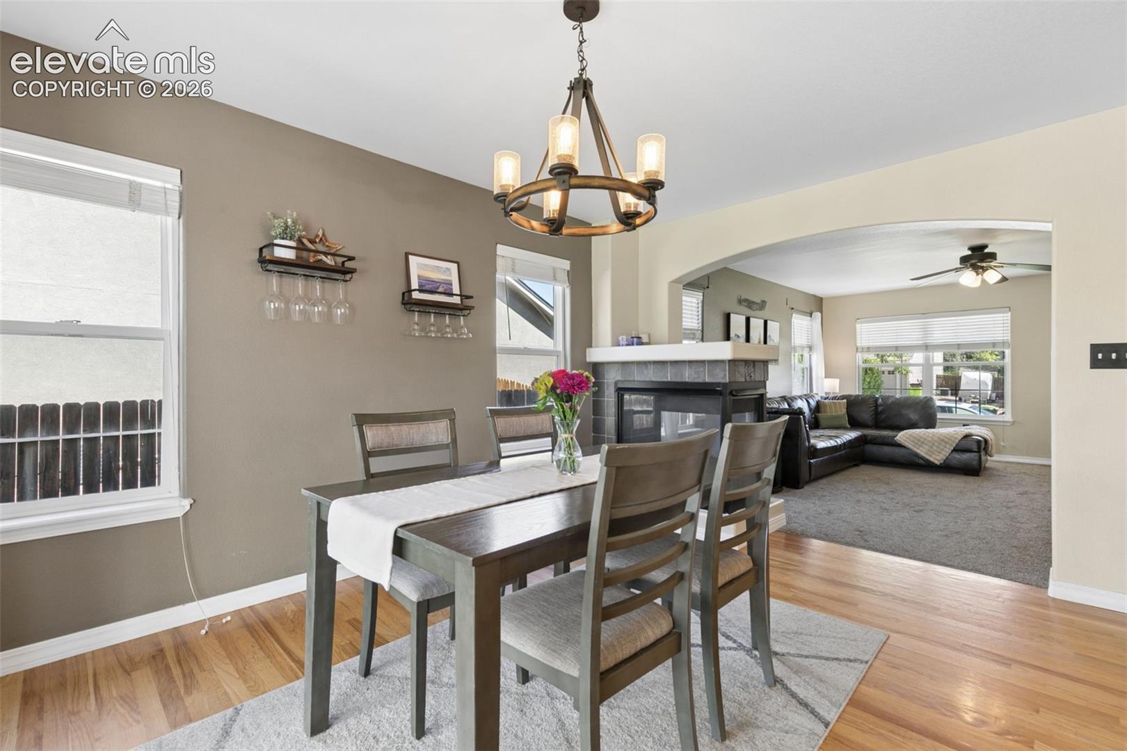 Wood-finish flooring, recessed windows with blinds, a multi-light chandelier, and an arched entry to a carpeted room featuring a ceiling fan and a fireplace with a stone surround