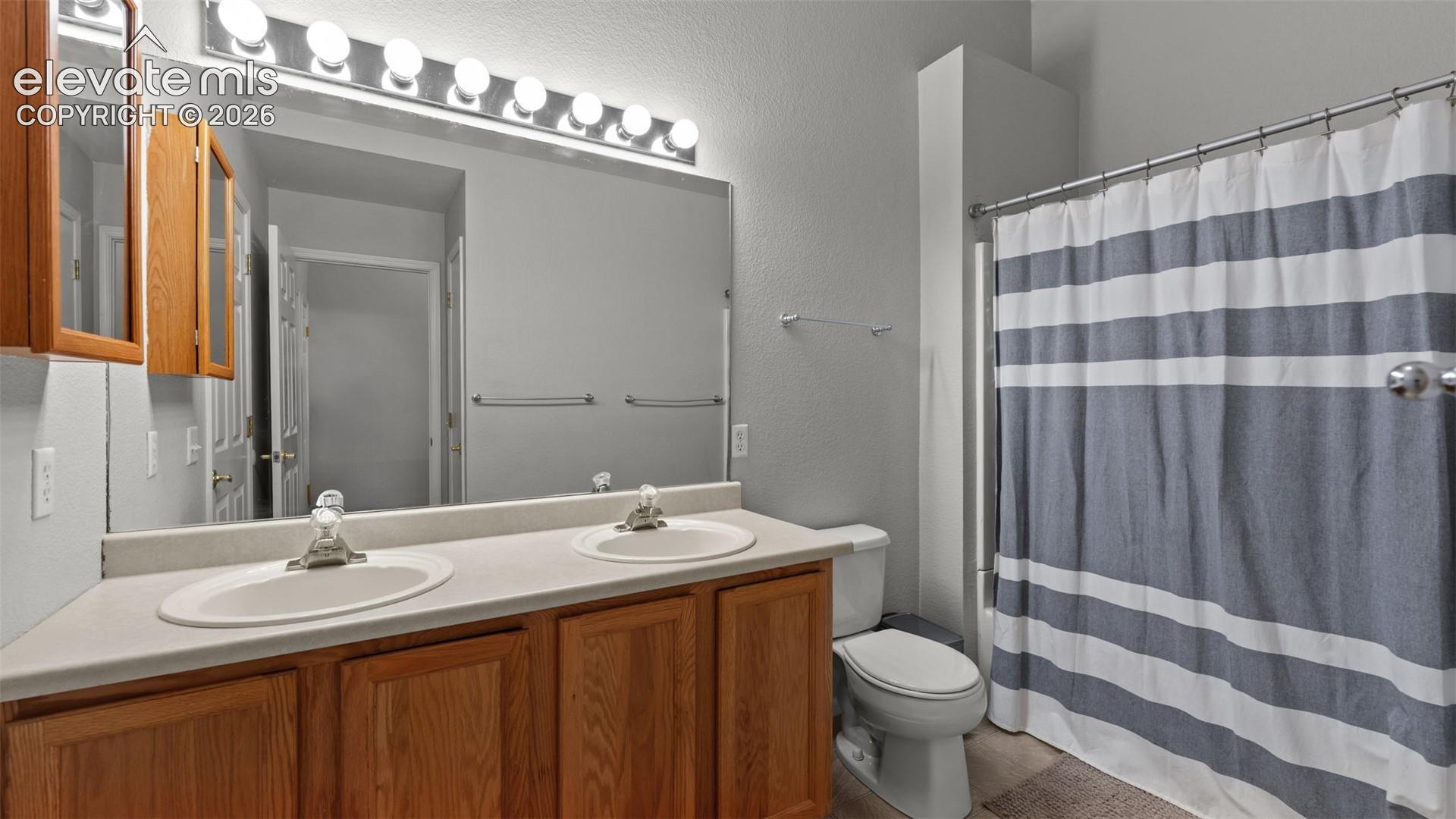 Bathroom featuring a dual-sink vanity with wood cabinetry, a solid-surface countertop, an expansive wall-mounted mirror, and an eight-bulb overhead light fixture