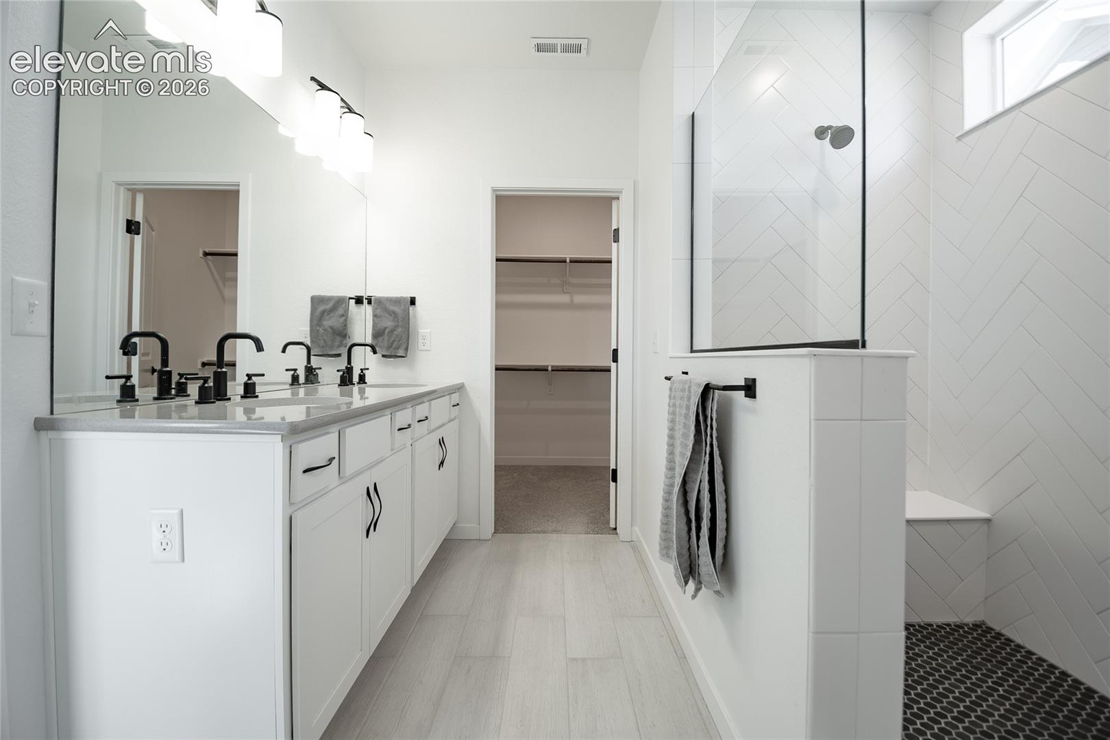 Dual-sink vanity with stone countertop and matte black fixtures, full-width mirror, and walk-in shower with white herringbone-patterned tile walls and integrated bench