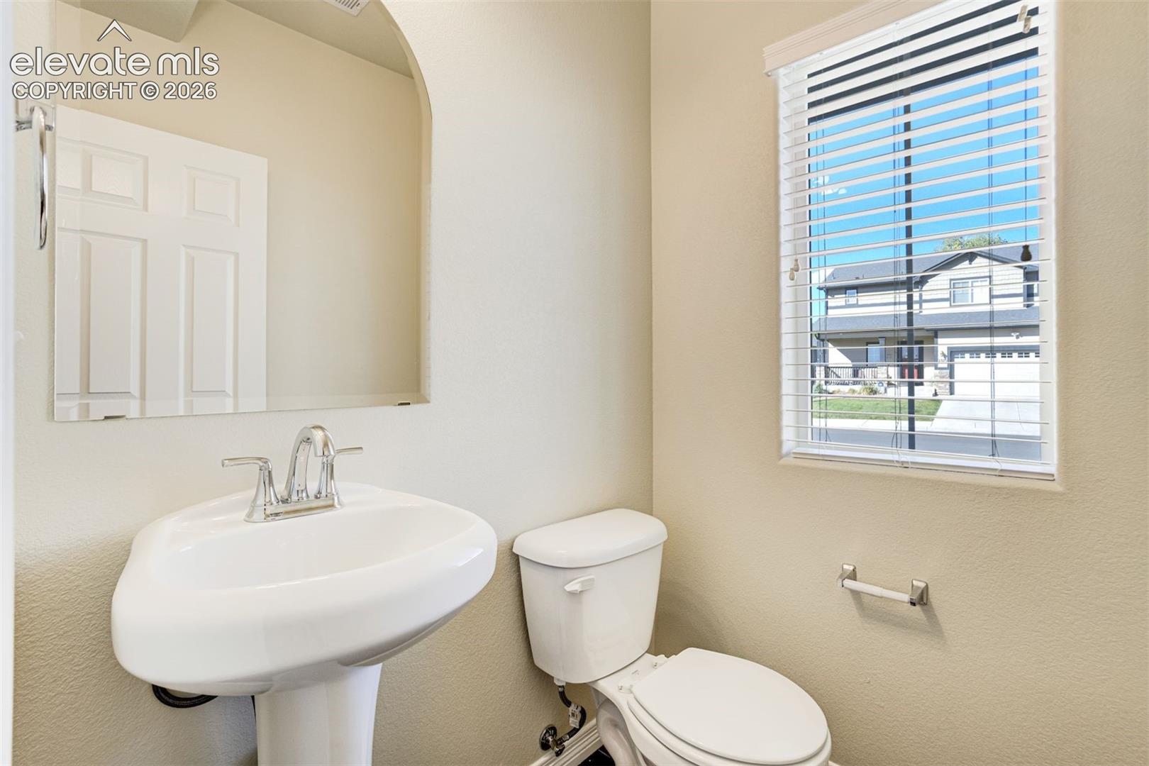 Pedestal sink with chrome faucet, wall-mounted mirror, white window blinds, and a white toilet in the main level powder bath