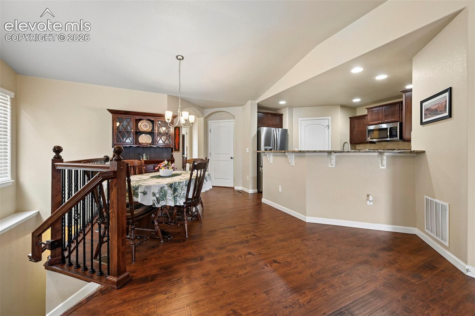 Open-concept living space vinyl flooring,  kitchen with dark wood cabinetry and stainless steel appliances, a breakfast bar with a speckled countertop, a dining area with a chandelier, and a wood staircase with wrought iron balusters